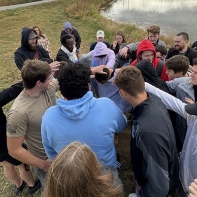 College students in a circle praying over a friend by a pond under an overcast sky.