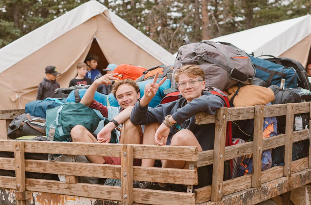 Two boys sit in the back of a wooden bed truck full of camping gear, smiling at the camera