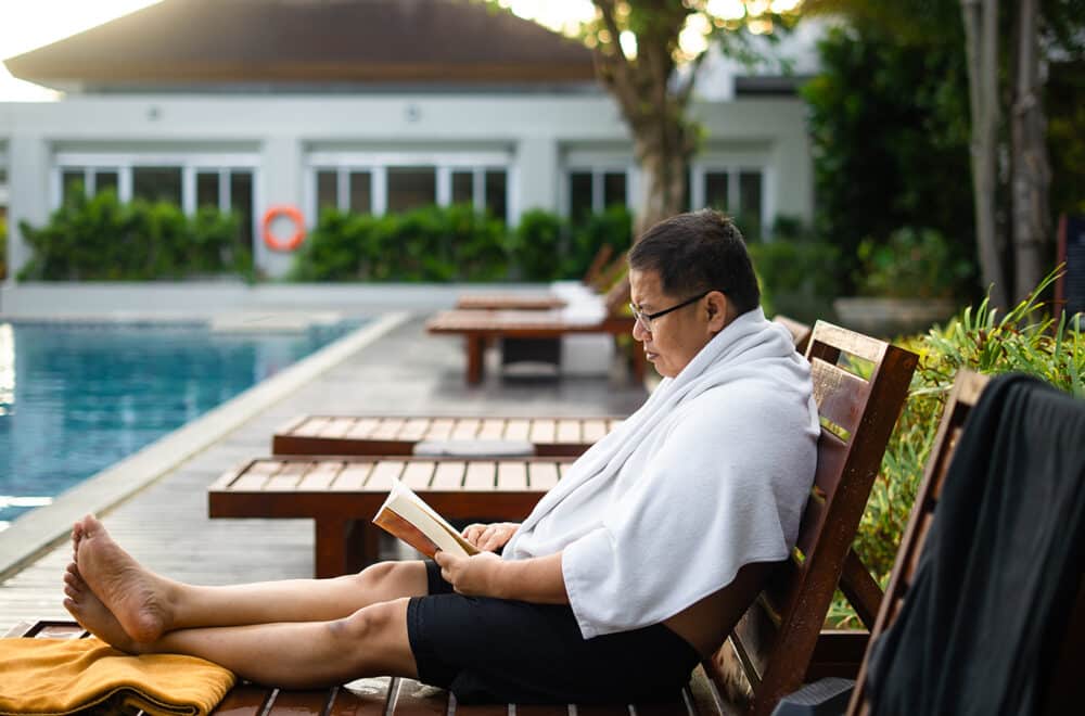 A man relaxes on a poolside lounge chair, wrapped in a white towel, reading a book with focused attention at a serene resort setting.