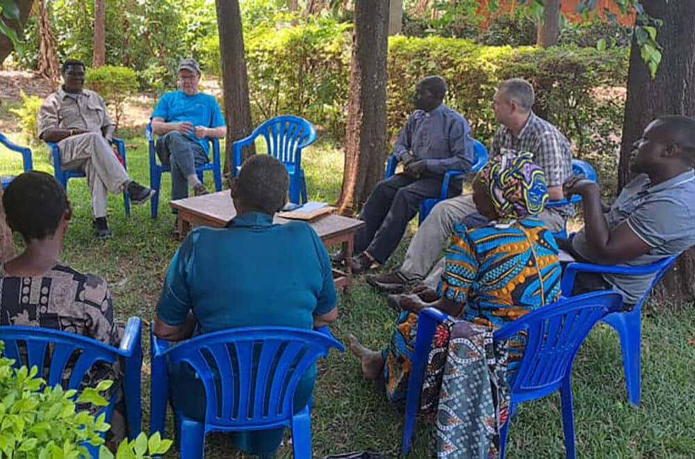 A diverse group of people gather in a circle of blue chairs under trees for a discussion, with a Bible open on a small wooden table in the center.