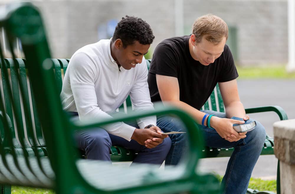 Two young men sit on a bench with Bibles in hand praying together.